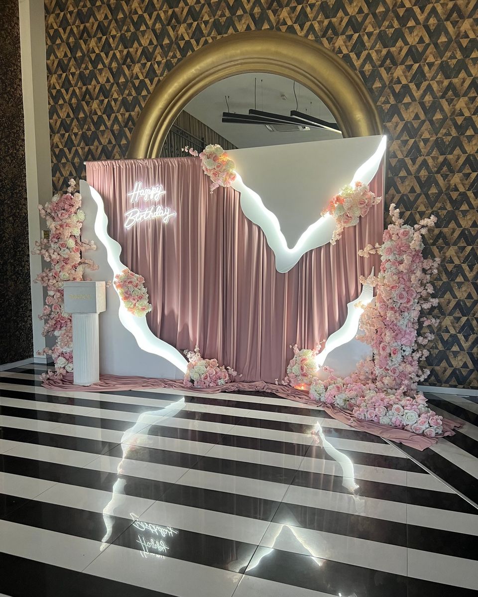 Roze draperija, roze cveće i beli talasasti panel sa natpisom Happy Birthday pred zlatnim ogledalom