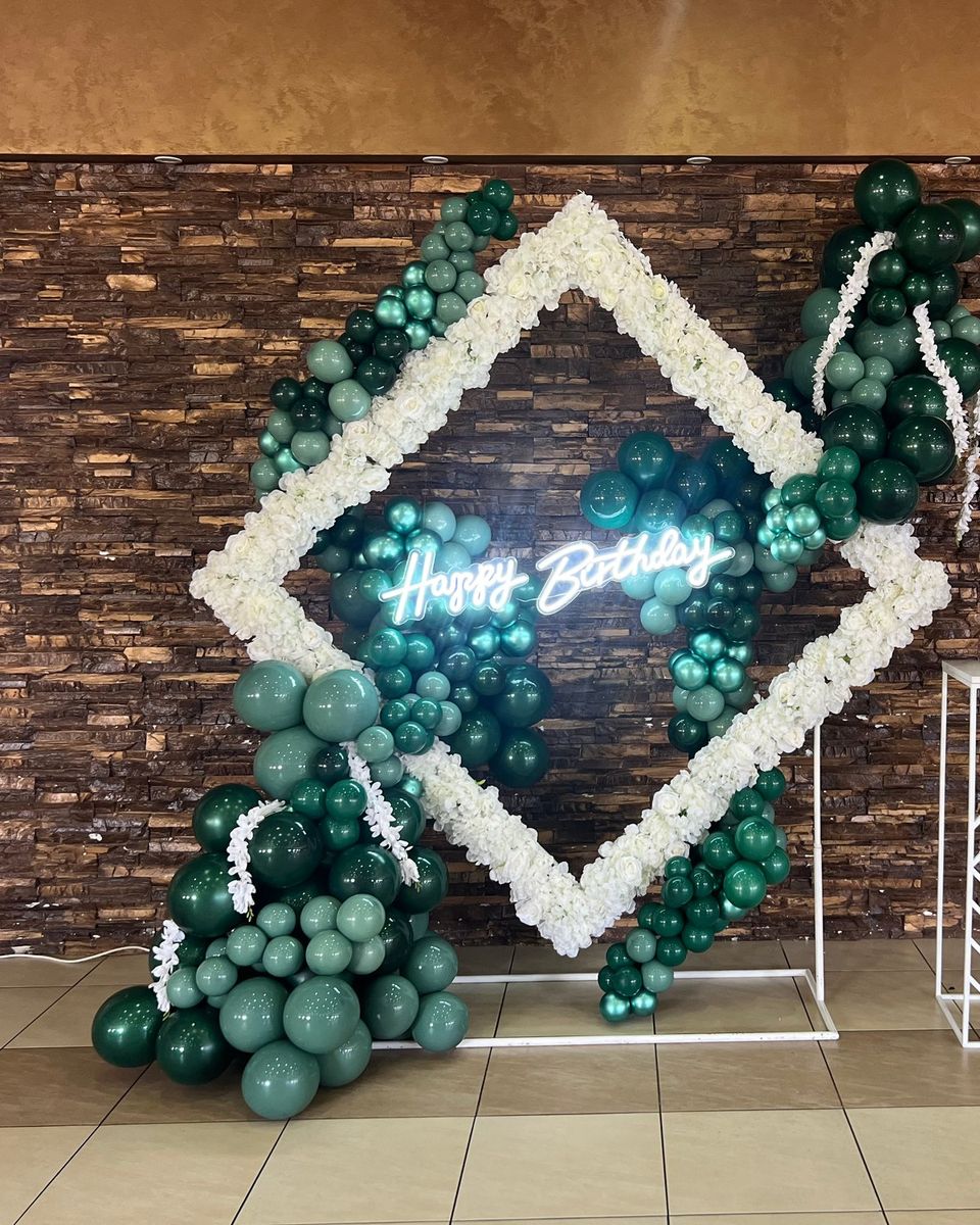 Beli cvetni romb sa smaragdno zelenim balonima i svetlećim natpisom uz zid od cigle