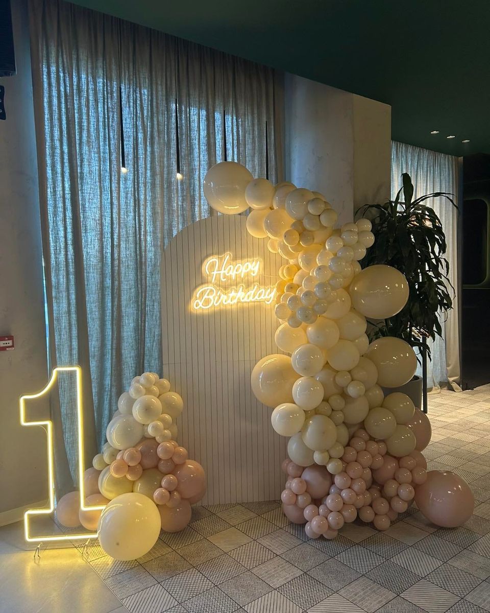 Krem, bež i roze baloni oko belog panoa sa natpisom Happy Birthday i svetlećim brojem 1