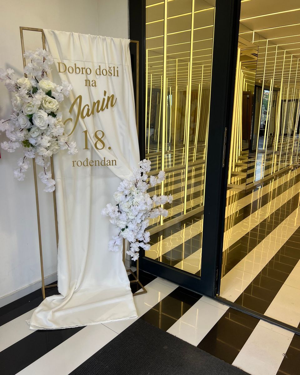 Beli satenski baner sa zlatnim slovima i belim cvećem za 18. rođendan pored ogledala