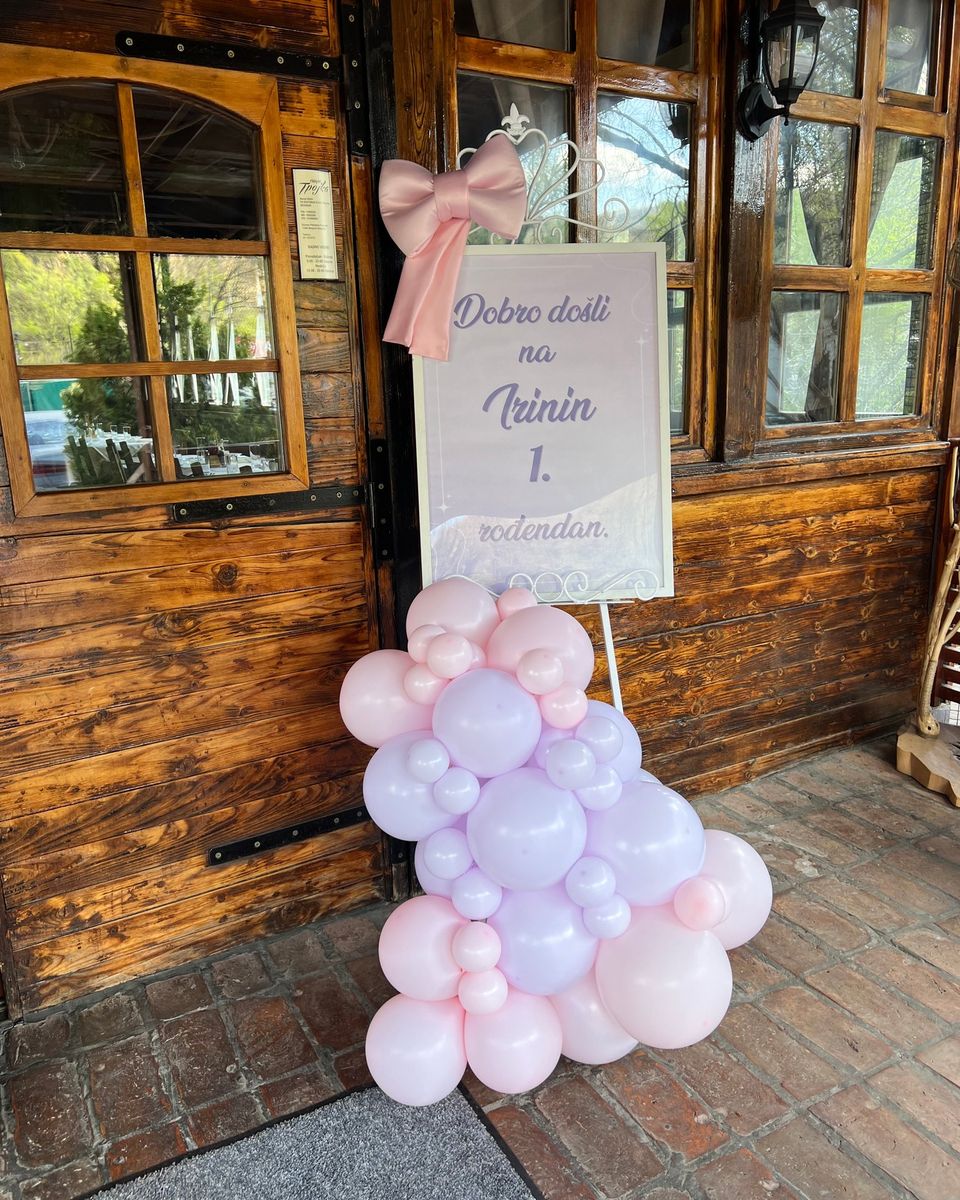 Tabla za prvi rođendan sa roze mašnom i lila i roze balonima ispred drvenih vrata