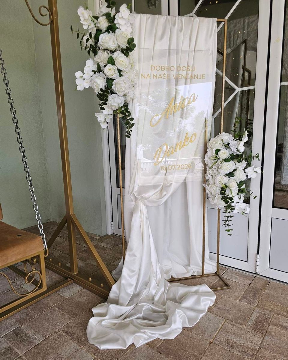 Tabla dobrodošlice za venčanje od bele draperije sa zlatnim slovima i belim cvećem