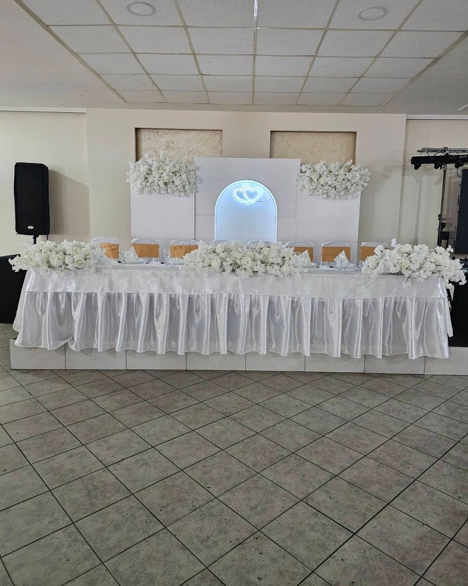 Beli glavni sto sa belim ružama ispred osvetljenog luka i aranžmana belog cveća