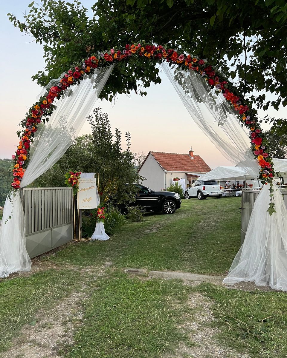 Luk od crvenog i narandžastog cveća sa belim tilom na ulazu, iza tabla dobrodošlice