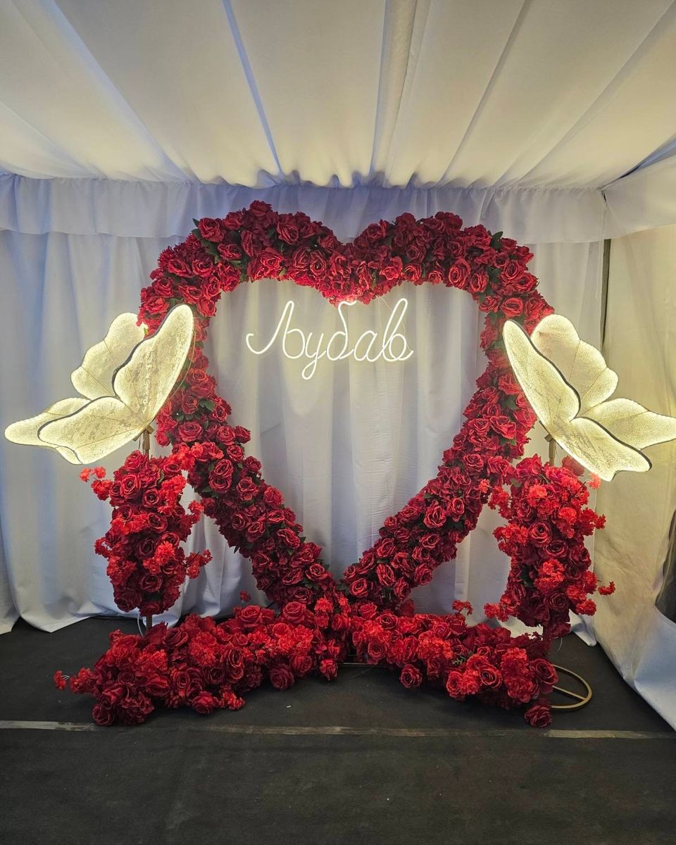 Veliko srce od crvenih ruža sa svetlećim leptirima i natpisom Ljubav ispred bele draperije